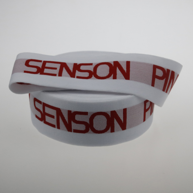 Two loops of white jacquard elastic tape with red SENSON PINKY text, stacked on a neutral studio surface