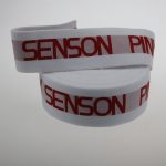 Folded white jacquard elastic strap with red SENSON PINKY text, resting on a light grey background