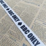 White ribbon with black BBC ONLY text and spade graphic, angled across a newspaper background