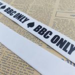 Two white woven straps with black BBC ONLY and spade prints, arranged diagonally across newspaper pages