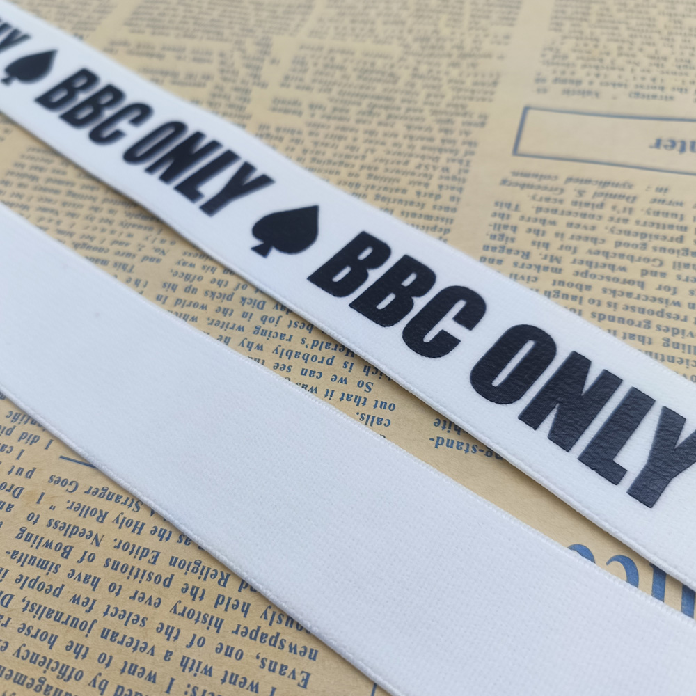 Two white woven straps with black BBC ONLY and spade prints, arranged diagonally across newspaper pages