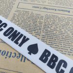 White woven ribbon with black BBC ONLY lettering and a spade motif, laid diagonally over vintage newspaper pages