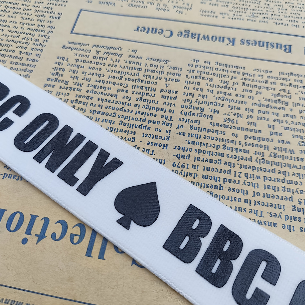 White woven ribbon with black BBC ONLY lettering and a spade motif, laid diagonally over vintage newspaper pages