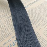 Angled close-up of navy blue herringbone twill webbing edge on newspaper, showing the woven selvedge detail