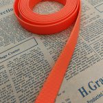 Coiled orange PVC-coated webbing strap with honeycomb embossed texture on newspaper, one end extending toward the corner