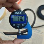 Digital thickness gauge with blue frame clamped onto a black honeycomb-textured webbing strap, reading 1.10 mm on the display