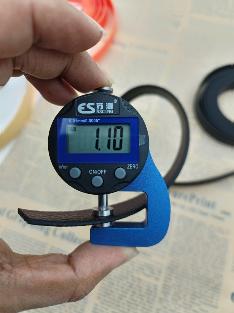 Digital thickness gauge with blue frame clamped onto a black honeycomb-textured webbing strap, reading 1.10 mm on the display