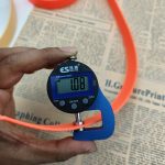 Digital thickness gauge with blue frame measuring an orange PVC-coated webbing strap, display reading 1.08 mm