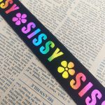 Black elastic band with rainbow laser-printed SISSY lettering and flower motifs laid on newspaper