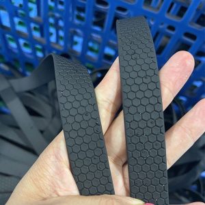 Two black PVC-coated webbing straps with raised honeycomb texture held in hand, water droplets on the surface, blue plastic crate behind
